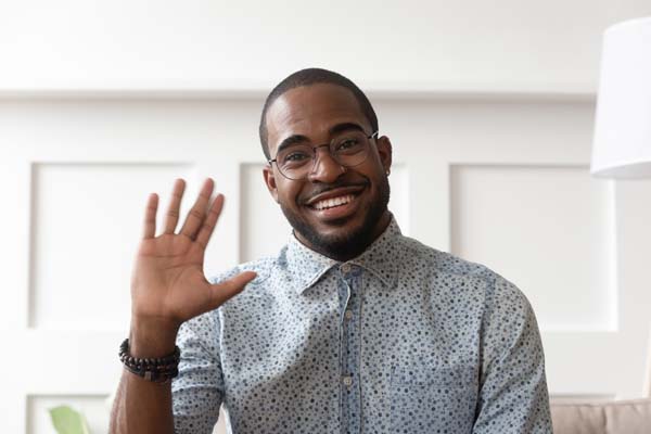 Smiling african American millennial man in glasses look at camera waving saying hello talking on video call, happy black male vlogger in spectacles greeting with subscribers shooting video blog