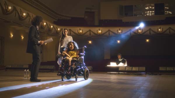 Actress with a disability driving around the stage in an electric wheelchair and rehearsing a performance with actors and a director in a dim theater, with a spotlight